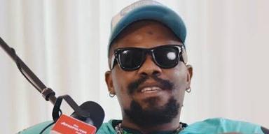 Rapper Ycee wearing a cap and sunglasses speaking into a microphone on the Afropolitan Podcast